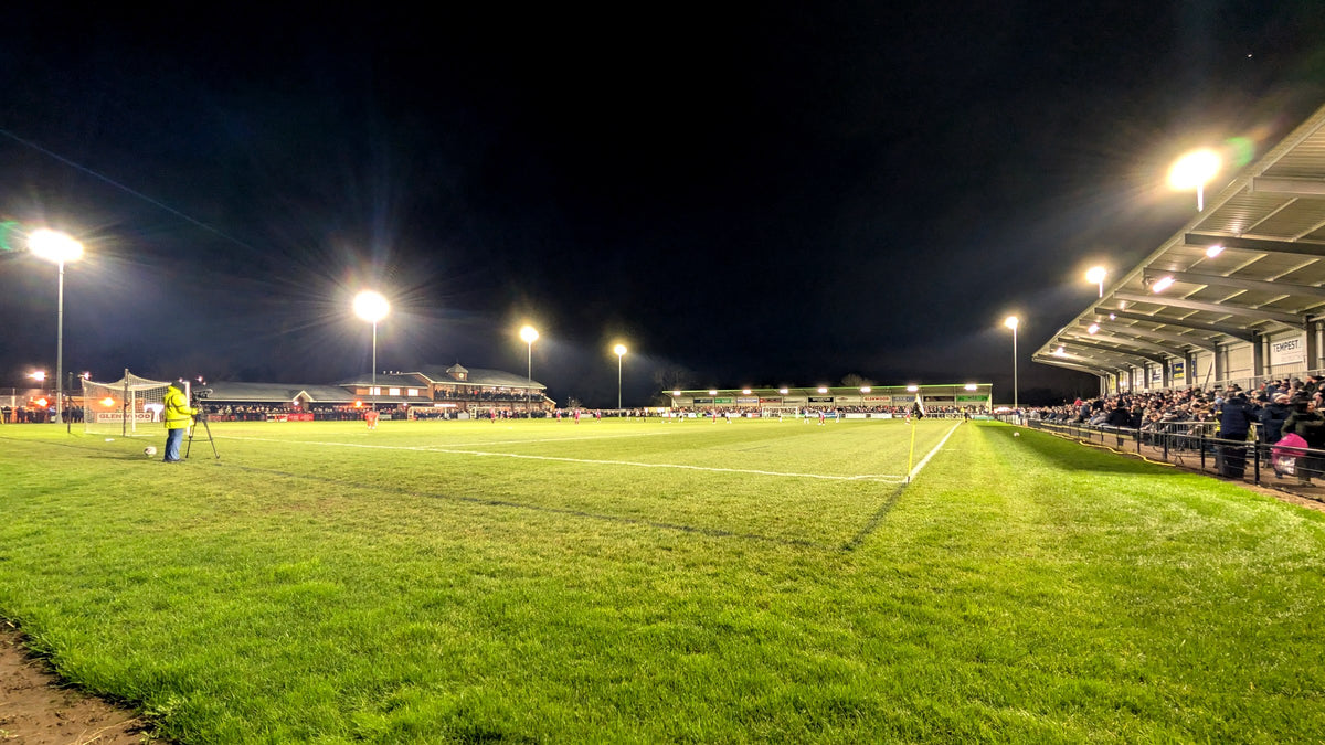 Floodlights over a non-league pitch at night, supporters along the touchline